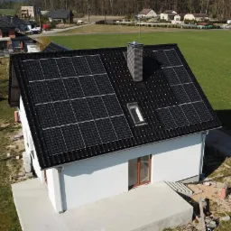 Widok z góry na dom jednorodzinny z czarnym dachem pokrytym panelami słonecznymi, komin z cegły i okno dachowe, na tle zielonego trawnika i zabudowy mieszkalnej.