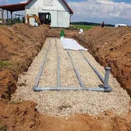 Drenaż rozsączający w wykopie wypełnionym żwirem, widoczne rury drenażowe, geowłóknina, w tle maszyna budowlana i budynek gospodarczy.