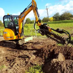 Żółta koparka JCB 8025 ZTS z zamontowaną frezarką łańcuchową wykonuje wykop w ziemi na tle zielonej trawy i drzew.