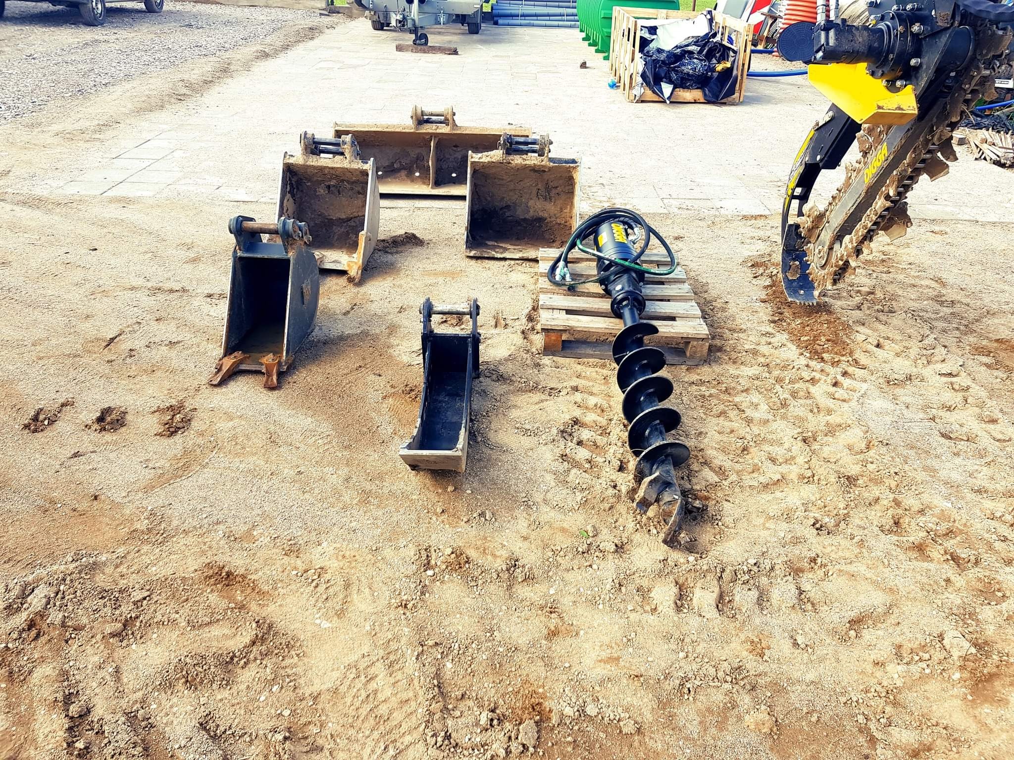 Różne łyżki koparkowe i wiertnica hydrauliczna leżące na piaszczystym placu budowy, obok widoczny fragment koparki łańcuchowej.