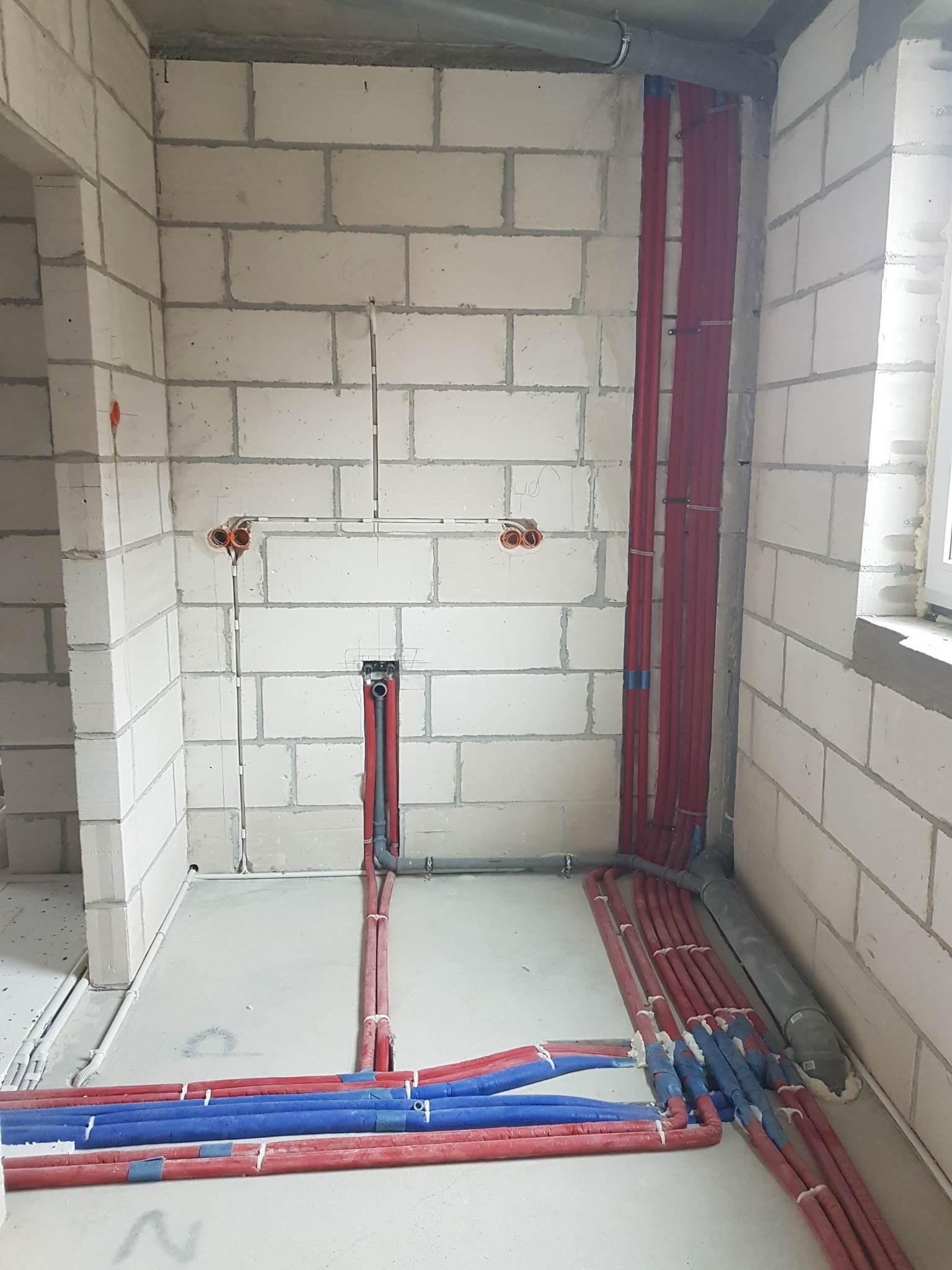 Surowy stan instalacji wodnej i centralnego ogrzewania w budynku z pustaków, widoczne rury czerwone i niebieskie, szare rury kanalizacyjne, gniazdka elektryczne w ścianie z betonu komórkowego.