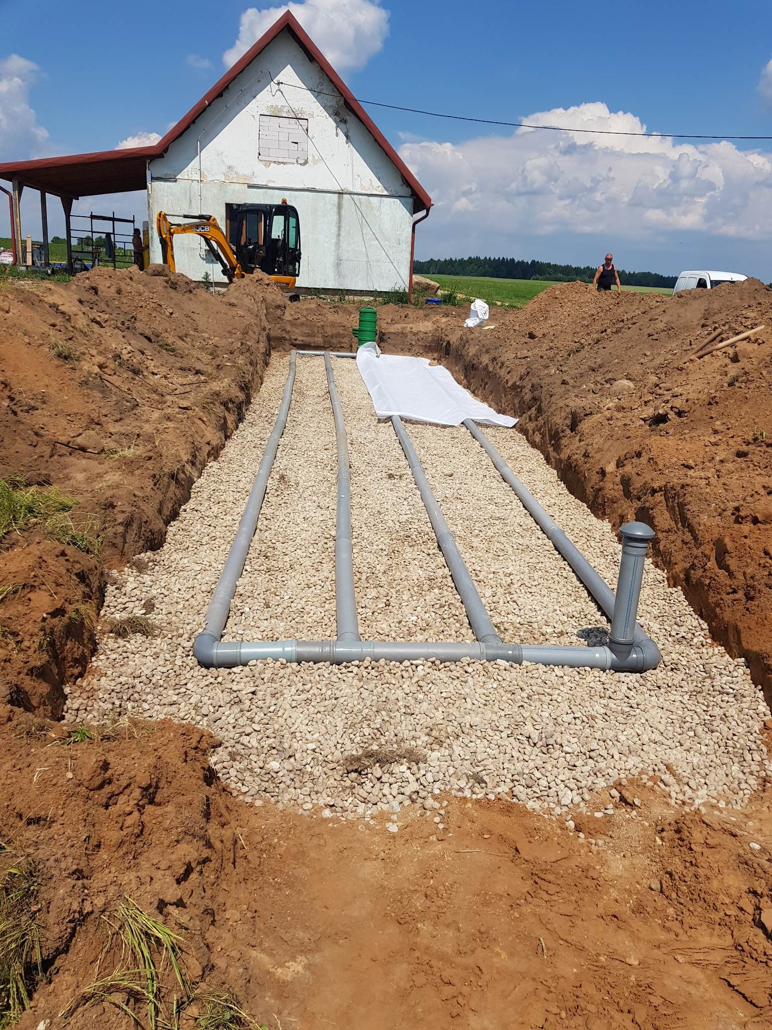 Drenaż rozsączający w wykopie wypełnionym żwirem, widoczne rury drenażowe, geowłóknina, w tle maszyna budowlana i budynek gospodarczy.