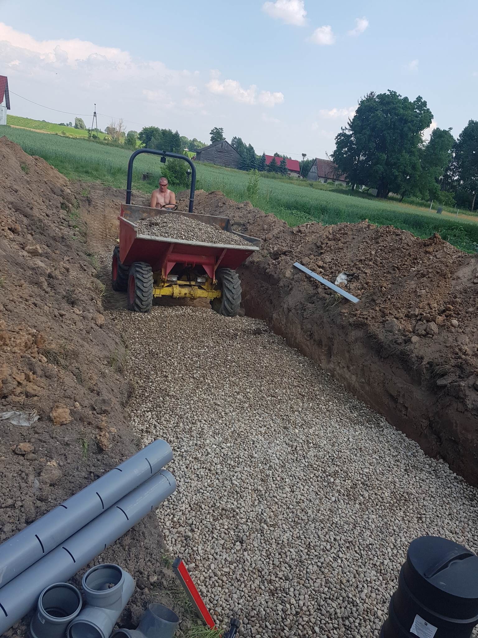 Wykop z drenażem żwirowym, widoczne rury drenażowe i pracownik na mini-wywrotce z żwirem na tle wiejskiego krajobrazu.