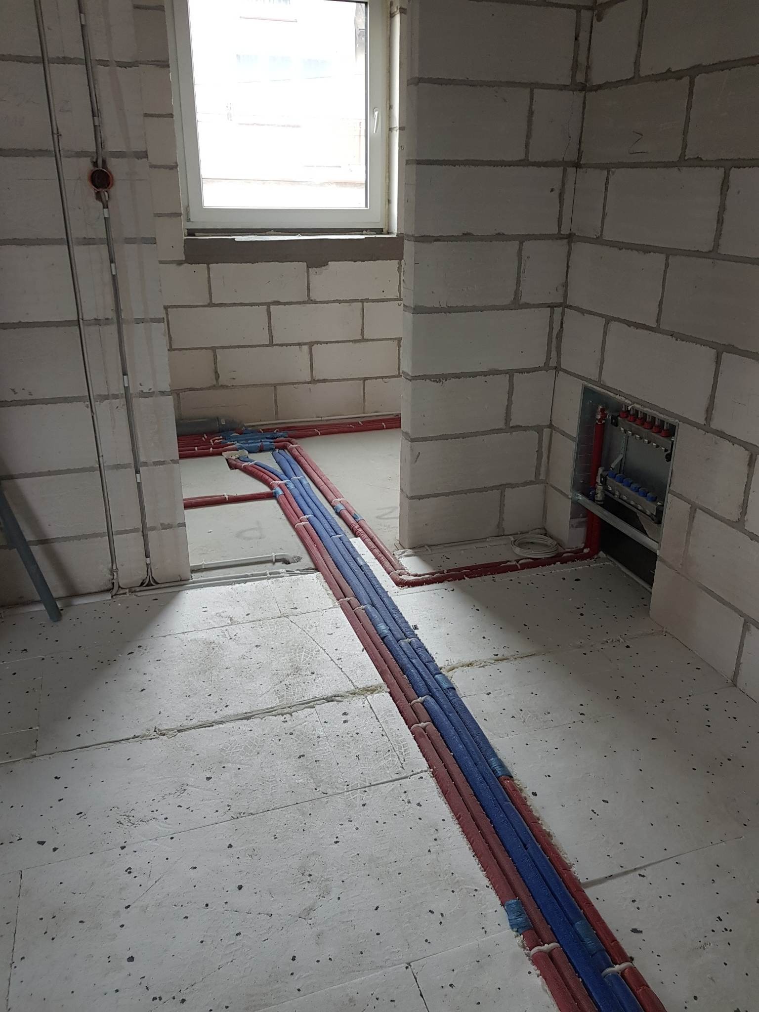 Surowy stan: rury grzewcze w izolacji czerwonej i niebieskiej ułożone na podłodze w nowym budownictwie, widoczny rozdzielacz w ścianie z pustaków, okno w tle.