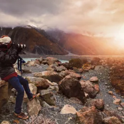 Fotograf siedzi na skale z aparatem na statywie, fotografując górski krajobraz z jeziorem i zachodzącym słońcem.