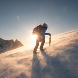 INNOVA SYSTEM Robert Przybyłek - Osoba z plecakiem i statywem wędruje po zasypanym śniegiem zboczu góry, pod słońce, widoczny silny wiatr i ośnieżone szczyty w tle.