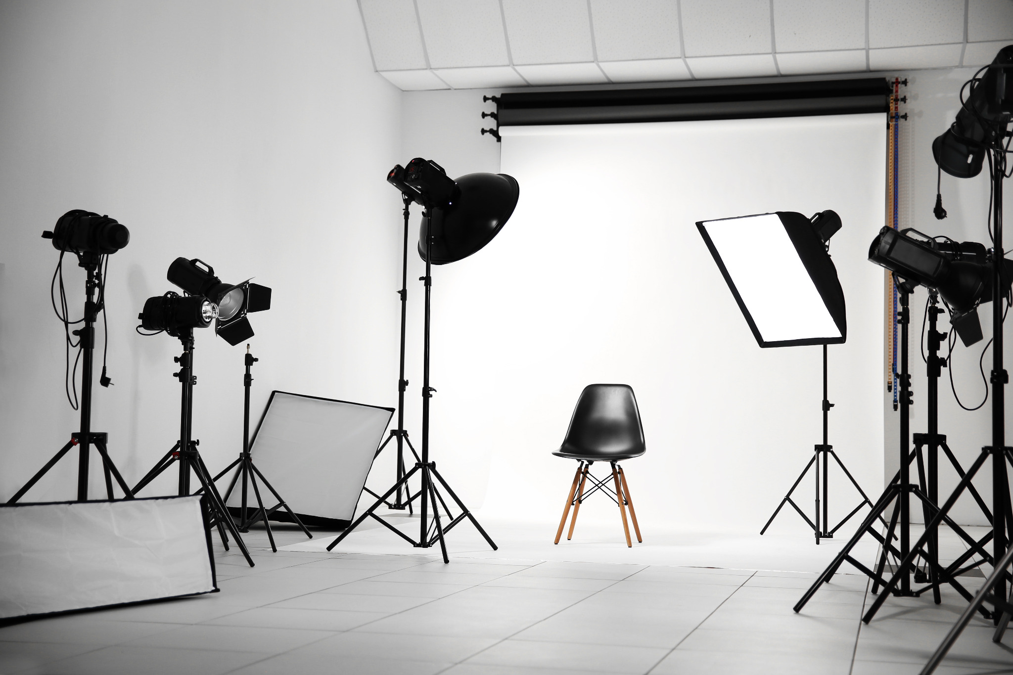 Profesjonalne studio fotograficzne z białym tłem, czarnym krzesłem Eames na drewnianych nogach i licznymi lampami studyjnymi z softboxami i reflektorami.
