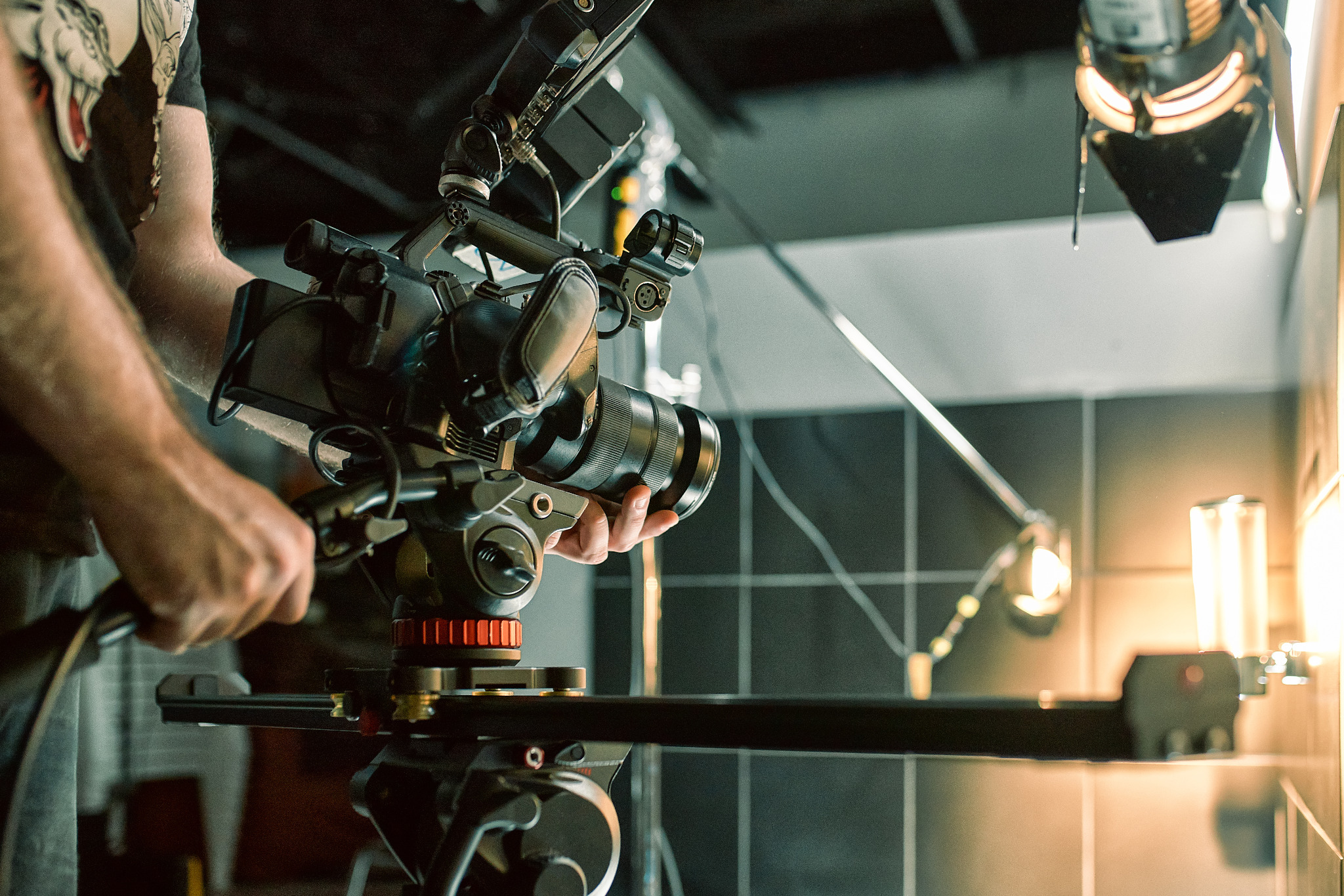 Profesjonalna kamera filmowa na statywie, z widocznymi rękami operatora, oświetlenie studyjne w tle, ujęcie z bliska.