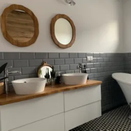 Nowoczesna łazienka z dwoma umywalkami nablatowymi, drewnianym blatem, szarą glazurą w stylu metro, lustrami w drewnianych ramach o nieregularnym kształcie i wolnostojącą wanną na ozdobnych nóżkach.