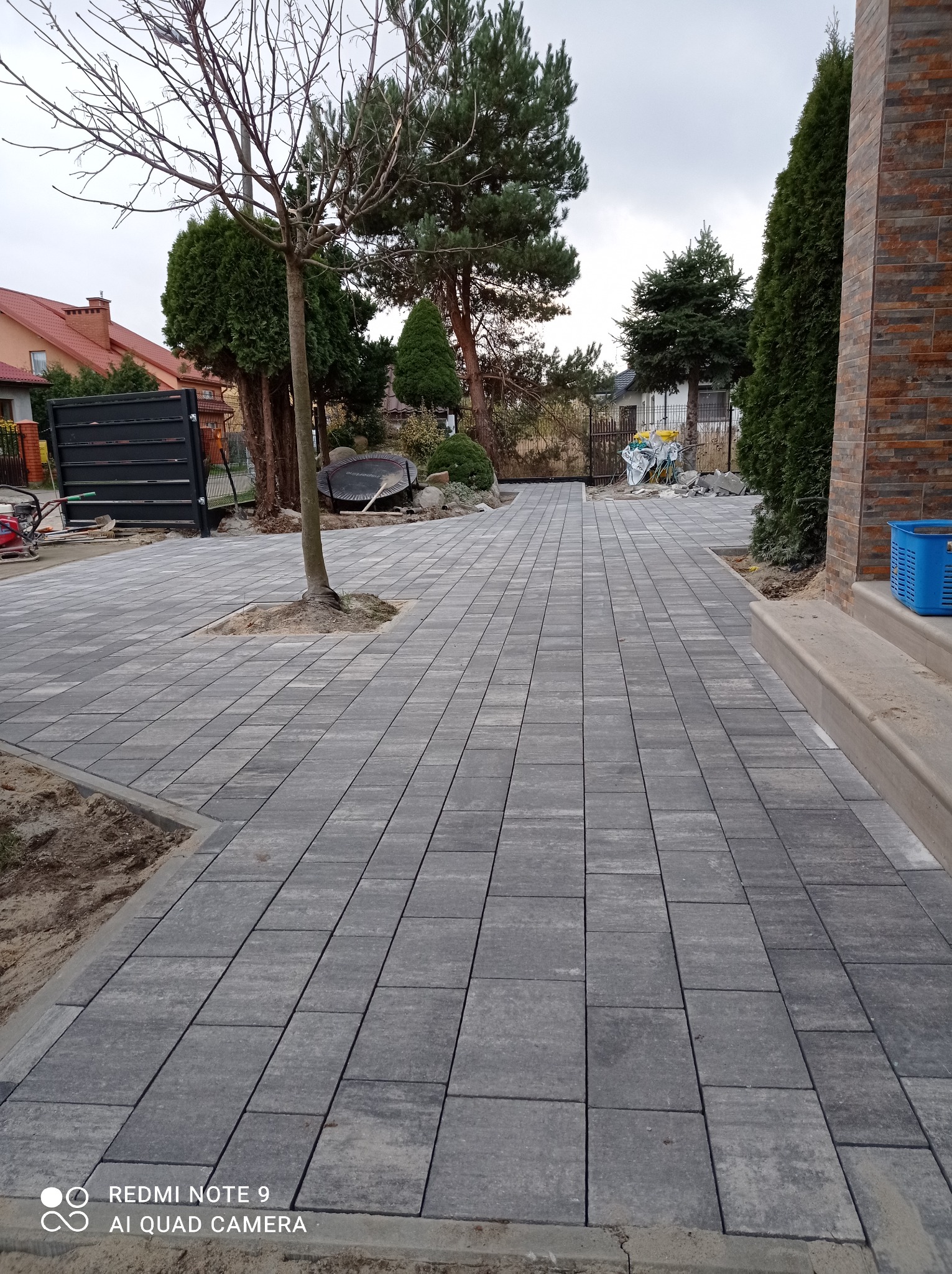 Ułożona szara kostka brukowa prowadząca do domu, z drzewem rosnącym w wycięciu w nawierzchni, widoczne narzędzia i materiały budowlane w tle.