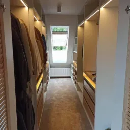 Wąska garderoba z białymi szafami wnękowymi oświetlonymi listwami LED, z oknem dachowym i beżową wykładziną podłogową.