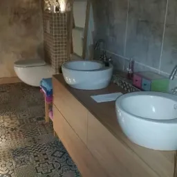 Rustykalna łazienka z dwoma okrągłymi umywalkami nablatowymi na drewnianej szafce, patchworkowa podłoga z płytek ceramicznych i ściany imitujące kamień.