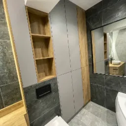 Nowoczesna łazienka z szarą zabudową meblową, drewnianymi akcentami i marmurowymi płytkami, widok na toaletę i odbicie umywalki w lustrze.