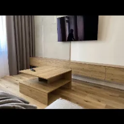 Nowoczesny salon z drewnianą szafką RTV i stolikiem kawowym z półkami, uzupełniony o szarą zasłonę i telewizor.