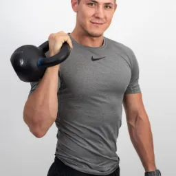 Uśmiechnięty mężczyzna w szarej koszulce trzyma czarny kettlebell z niebieskim akcentem na uchwycie, widoczny zegarek na nadgarstku.