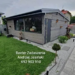 Nowoczesna, drewniana altana z dużym oknem i szarymi drzwiami, otoczona trawnikiem, kamieniami dekoracyjnymi i chodnikiem z kostki brukowej.