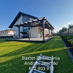 Nowoczesna pergola z ciemnego drewna nad betonowym tarasem, przylegająca do jasnego domu z dużymi oknami, widok na zadbany trawnik i ogrodzenie, drabina oparta o trawę w pierwszym planie.