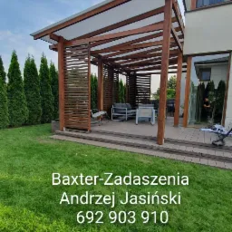 Drewniana pergola z częściowo zabudowanymi ścianami, zadaszona przezroczystym materiałem, stoi na tarasie z kompozytu, obok budynku z dużym oknem. Wewnątrz pergoli znajdują się meble ogrodowe.