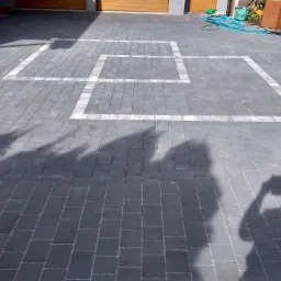 Szara kostka brukowa ułożona na podjeździe z białymi obramowaniami w kształcie kwadratów, cień osoby robiącej zdjęcie widoczny na powierzchni.