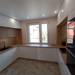 H&H Meble na wymiar - Nowoczesna kuchnia w kształcie litery U, białe fronty szafek z drewnianymi akcentami, czarny zlew i bateria, marmurowa podłoga, okno z widokiem na budynek z czerwonym dachem.