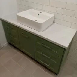 Łazienka z zieloną szafką pod umywalkę nablatową, biały blat z delikatnym wzorem, białe kafelki na ścianie i szare płytki na podłodze.