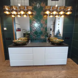 Łazienka z dwoma umywalkami w stylu glamour, zielone płytki heksagonalne na ścianie, biała szafka pod umywalki z szufladami, drewniana podłoga i złote lampy wiszące.