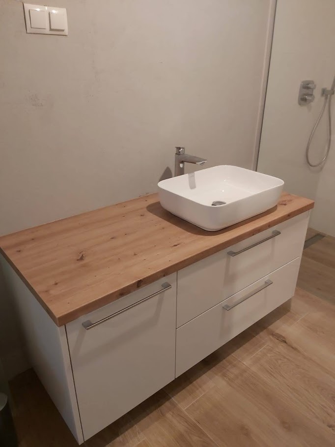 Łazienkowa szafka z białymi frontami i drewnianym blatem, na którym umieszczona jest kwadratowa umywalka nablatowa i nowoczesna bateria.