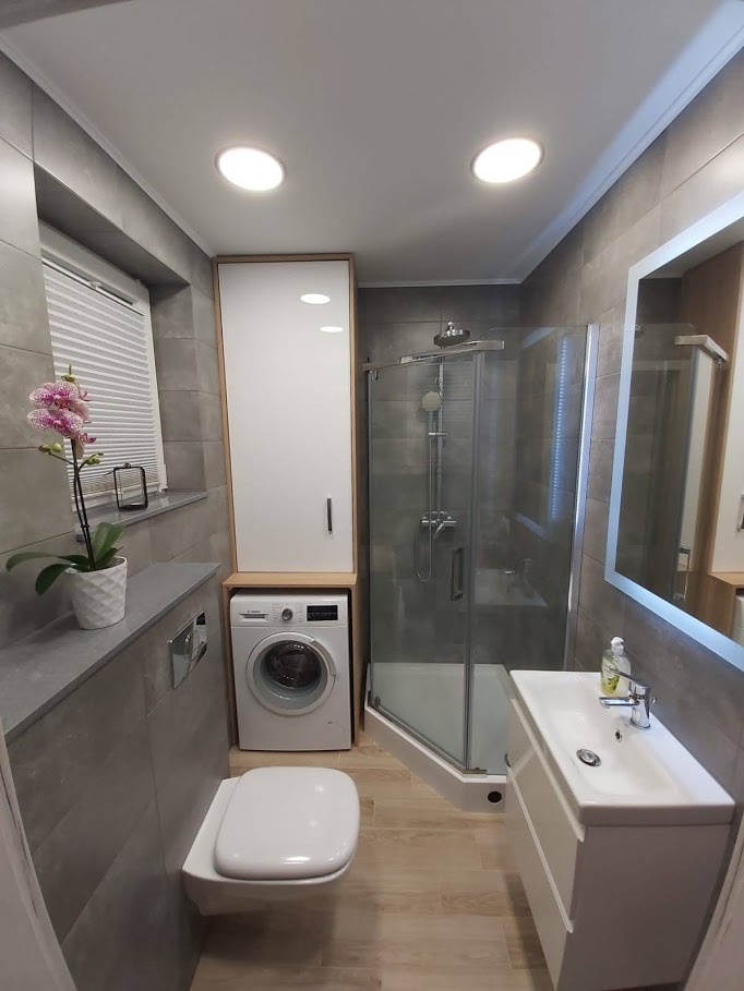 Nowoczesna łazienka z szarymi płytkami, kabiną prysznicową narożną, umywalką z szafką, toaletą podwieszaną i pralką w zabudowie z wysoką szafką, akcentem kolorystycznym jest storczyk na parapecie...