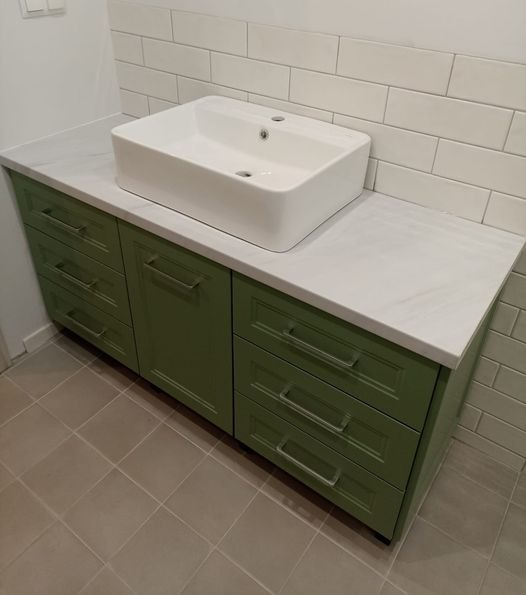 Łazienka z zieloną szafką pod umywalkę nablatową, biały blat z delikatnym wzorem, białe kafelki na ścianie i szare płytki na podłodze.