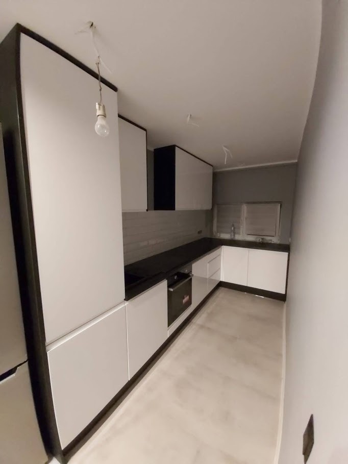 Nowoczesna kuchnia z białymi frontami szafek i czarnym blatem, widoczna żarówka na kablu sugerująca etap wykończeniowy, szare ściany i podłoga.