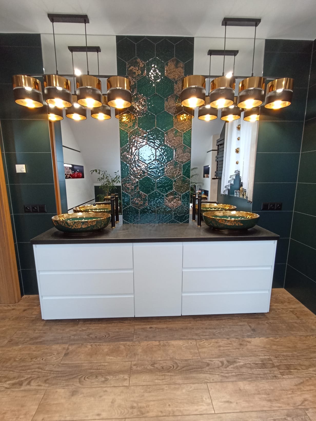 Łazienka z dwoma umywalkami w stylu glamour, zielone płytki heksagonalne na ścianie, biała szafka pod umywalki z szufladami, drewniana podłoga i złote lampy wiszące.
