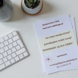 Biurko z minimalistyczną klawiaturą, wazonem i ulotką reklamową z hasłem 'Nie masz swojej strony? Potrzebujesz zrobić nową? Wykonamy ją dla Ciebie!' oraz danymi kontaktowymi Wydawnictwa OMA.