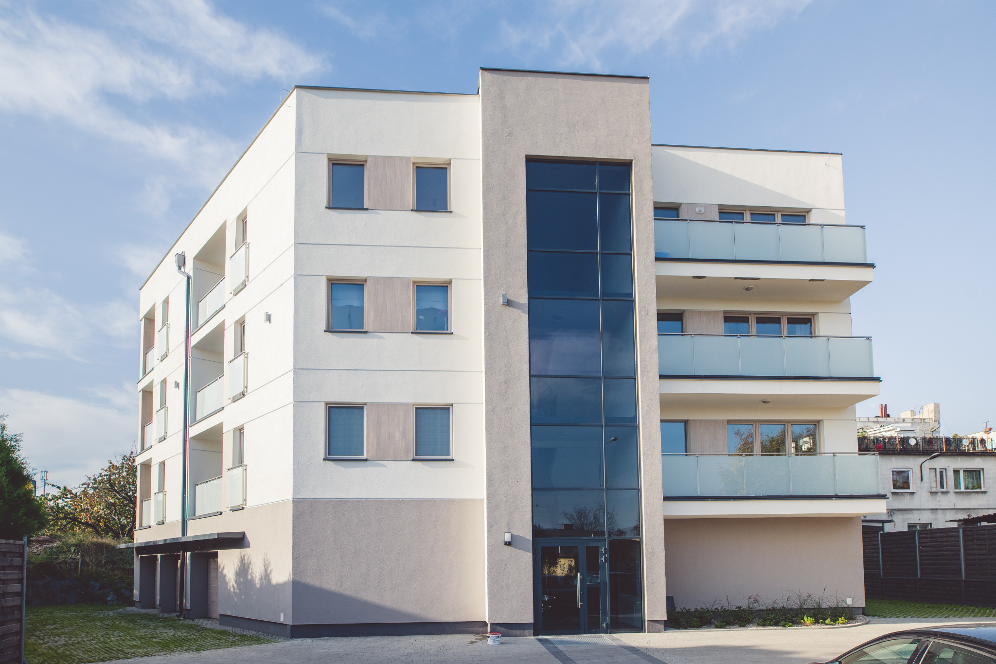Nowoczesny, trzypiętrowy budynek mieszkalny z balkonami i dużymi oknami, wykończony w bieli i szarości, widziany z perspektywy ulicy w słoneczny dzień.