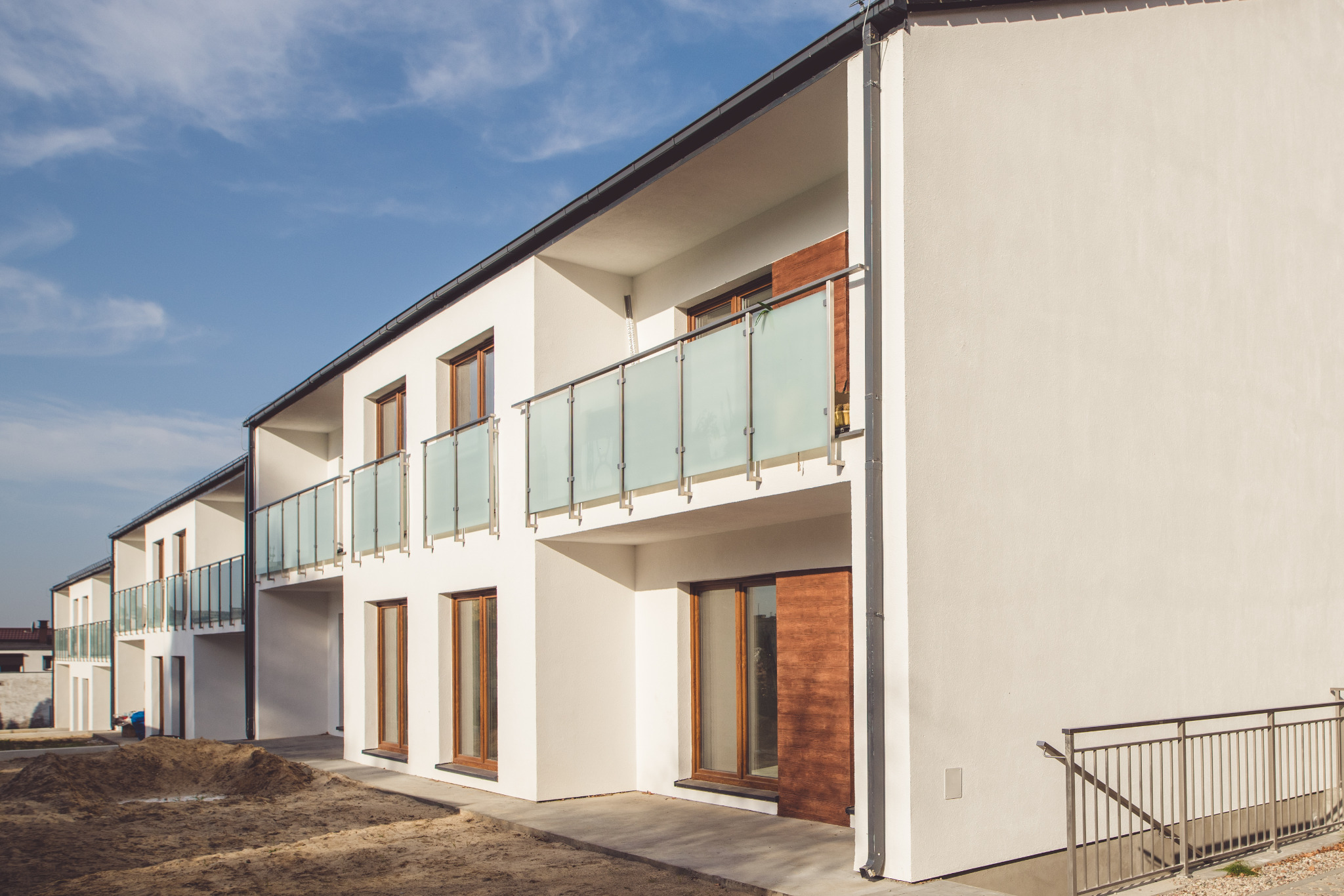 Rząd nowych, białych domów szeregowych z drewnianymi drzwiami i balkonami z matowego szkła, teren wokół w trakcie zagospodarowywania.