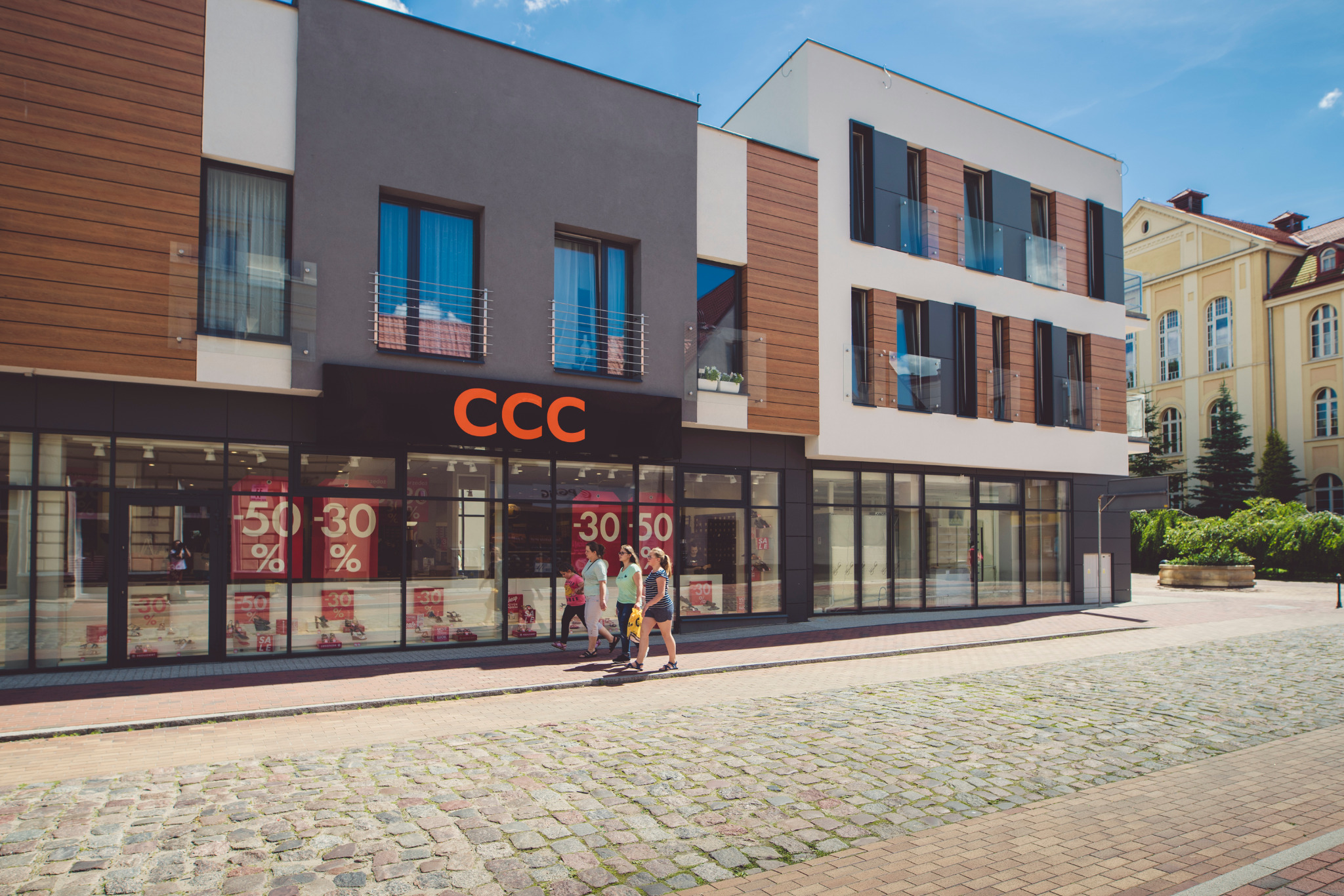 Nowoczesny budynek usługowy z elementami drewna i szarego tynku, z dużymi oknami wystawowymi i balkonami z balustradami ze stali nierdzewnej, na pierwszym planie brukowa ulica i przechodzące osoby.