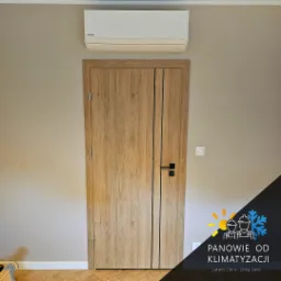 Klimatyzator Panasonic zamontowany nad drewnianymi drzwiami z czarną klamką, na tle beżowej ściany. Widoczne logo firmy oferującej usługi klimatyzacyjne.