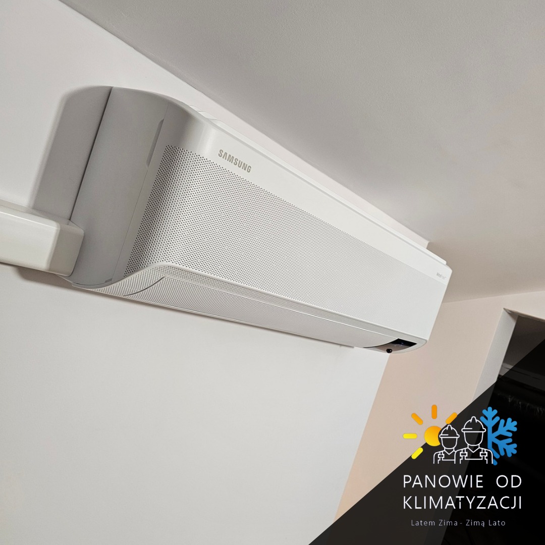 Biała klimatyzacja Samsung zamontowana na ścianie w jasnym pomieszczeniu. Widoczny fragment logo firmy monterskiej w prawym dolnym rogu.