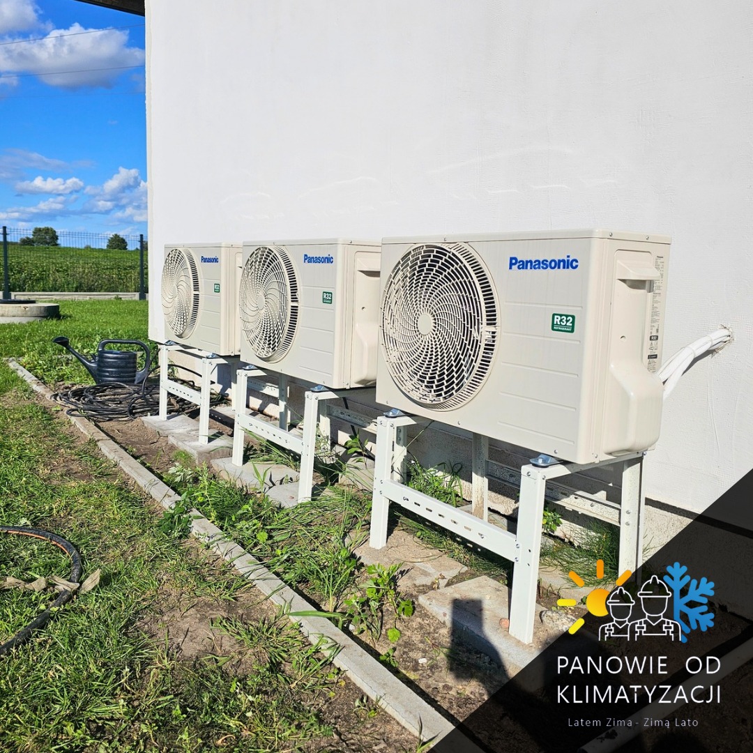 Trzy jednostki zewnętrzne klimatyzacji Panasonic R32 na metalowych wspornikach przy białej ścianie budynku. W tle zielony krajobraz i błękitne niebo.