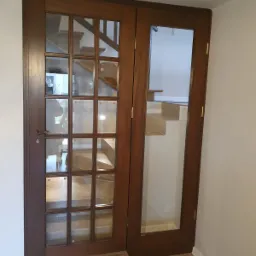 Dwuskrzydłowe, drewniane drzwi wewnętrzne z szybami w szprosach, widok na schody z płytek w tle.