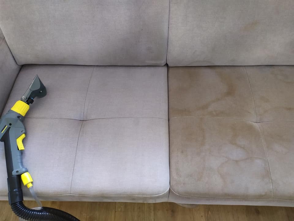 Czyszczenie jasnobeżowej kanapy z widocznymi plamami, na jednej połowie już wyczyszczonej, z użyciem odkurzacza piorącego Karcher z żółtymi elementami.