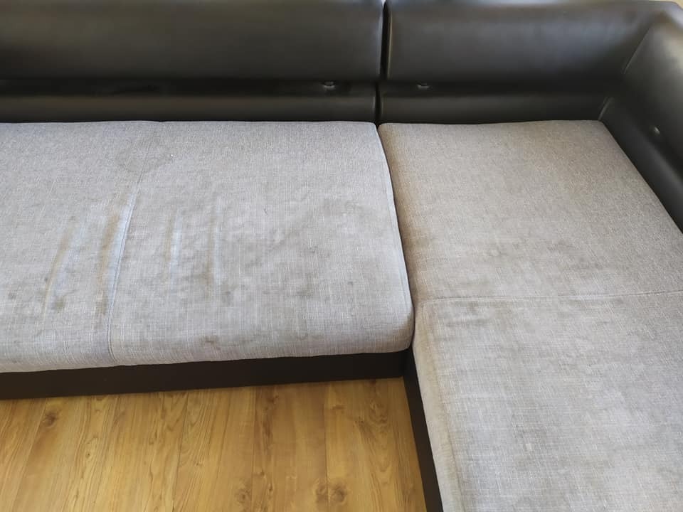 Fragment narożnika z szarą, zabrudzoną tapicerką na drewnianej podłodze, widoczne plamy i ślady użytkowania.