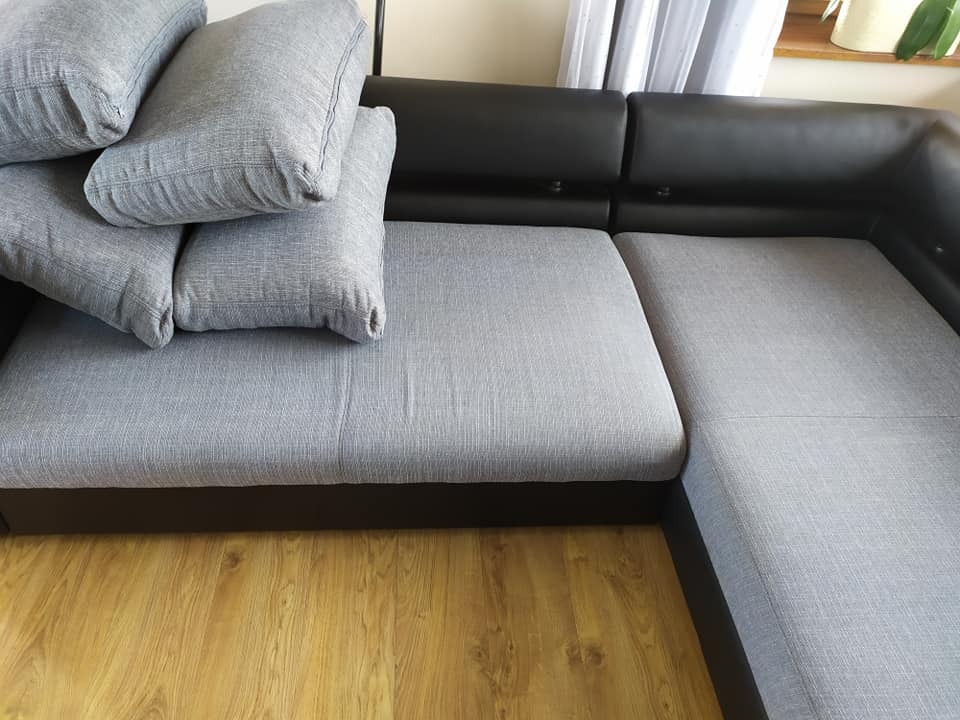 Szary narożnik z czarną ekoskórą i stertą poduszek w tym samym odcieniu szarości, leżący na drewnianej podłodze w jasnym pomieszczeniu.