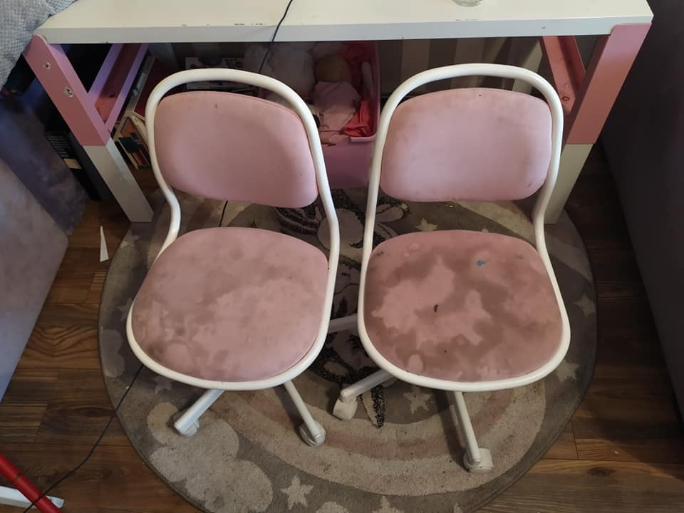 Dwa brudne, różowe krzesła biurowe z białymi ramami stoją obok siebie na wzorzystym dywanie, widoczne plamy na siedziskach, pod białym biurkiem z różowymi nogami.
