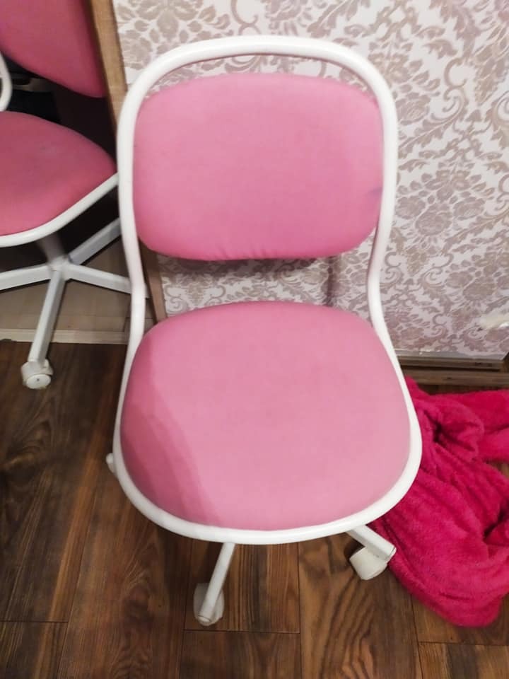 Różowe krzesło biurowe z białą ramą, widoczne plamy na siedzisku, na podłodze leży różowy koc, tło stanowi tapeta w kwiaty.