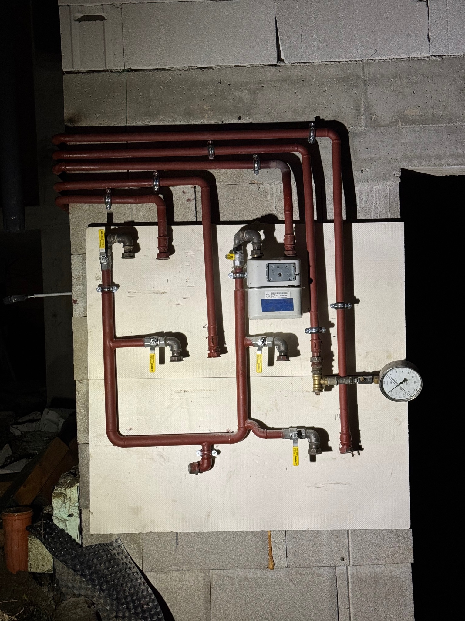 Czerwone rury gazowe z zaworami i manometrem zamontowane na białej płycie izolacyjnej na tle ściany z bloczków betonowych.