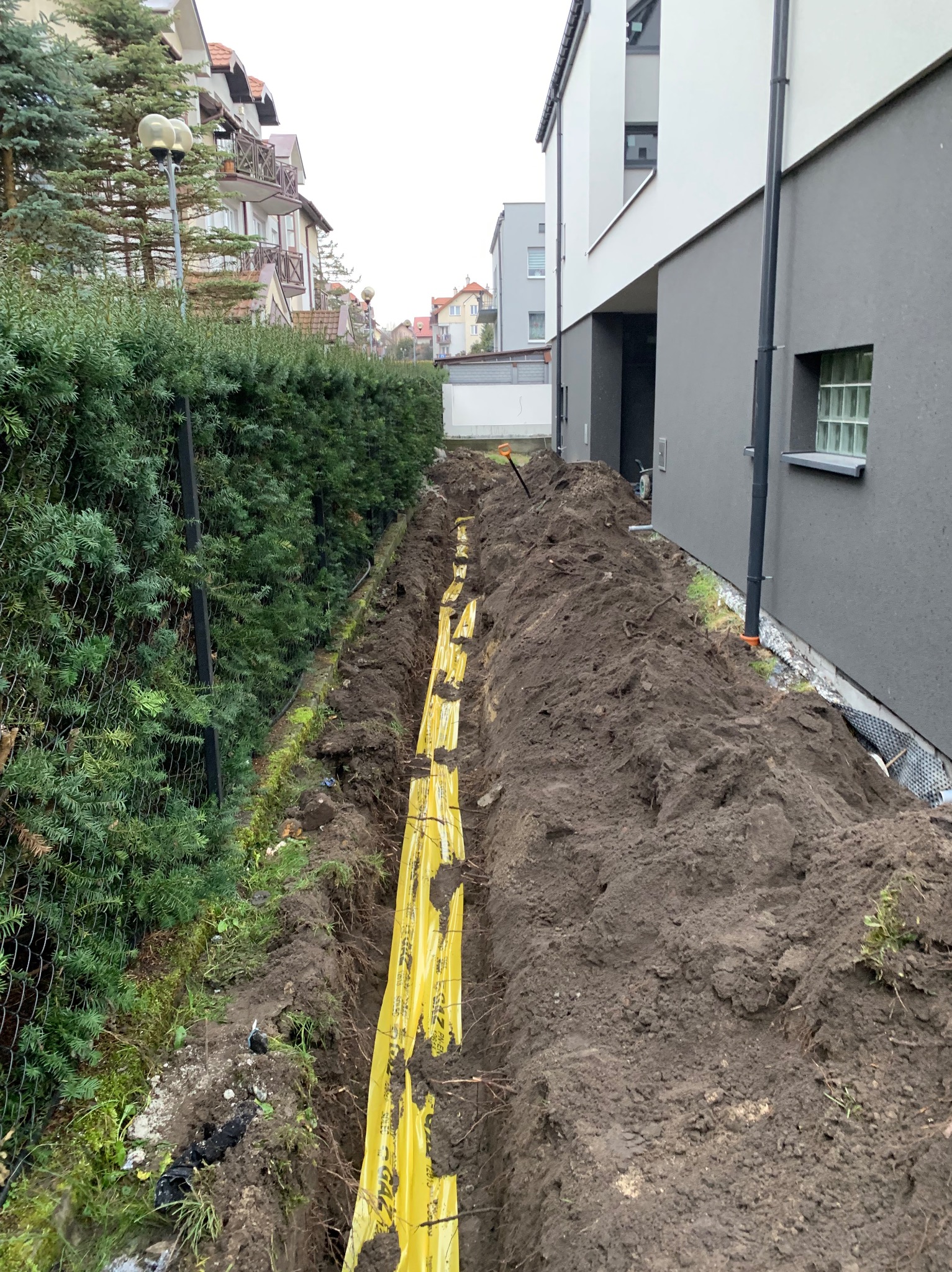 Wykop w ziemi z żółtą folią ostrzegawczą, prawdopodobnie pod instalację; widoczny fragment budynku i ogrodzenie z żywopłotu.