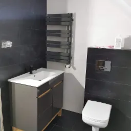 Nowoczesna łazienka w odcieniach szarości i bieli z wiszącą toaletą, grzejnikiem drabinkowym, szafką pod umywalkę z drewnianymi akcentami i czarną armaturą, widoczne niedokończone gniazdka...