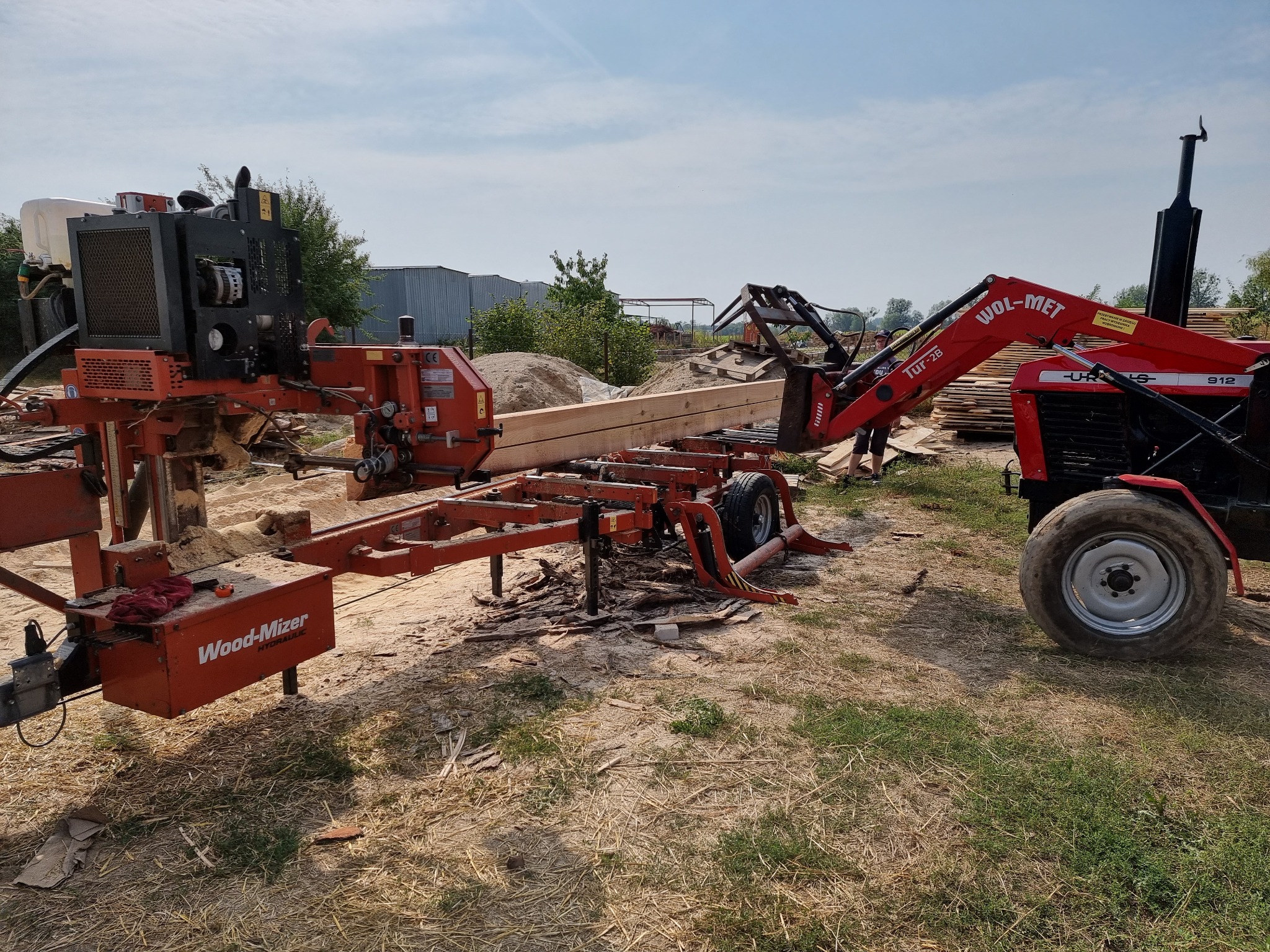 Mobilny tartak Wood-Mizer z podajnikiem hydraulicznym i traktorem Ursus 912 z przodu, przygotowany do cięcia długiej kantówki na placu tartacznym.