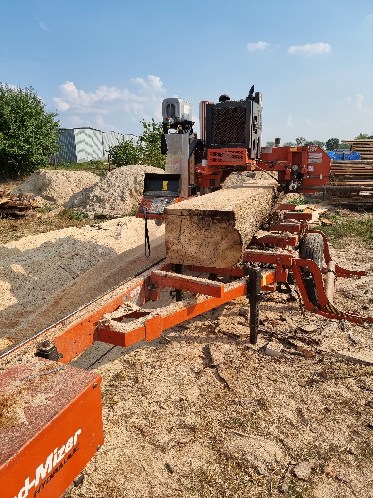 Pomarańczowa, przenośna traka tnie kłodę drewna na deski; w tle sterta desek i budynki gospodarcze.