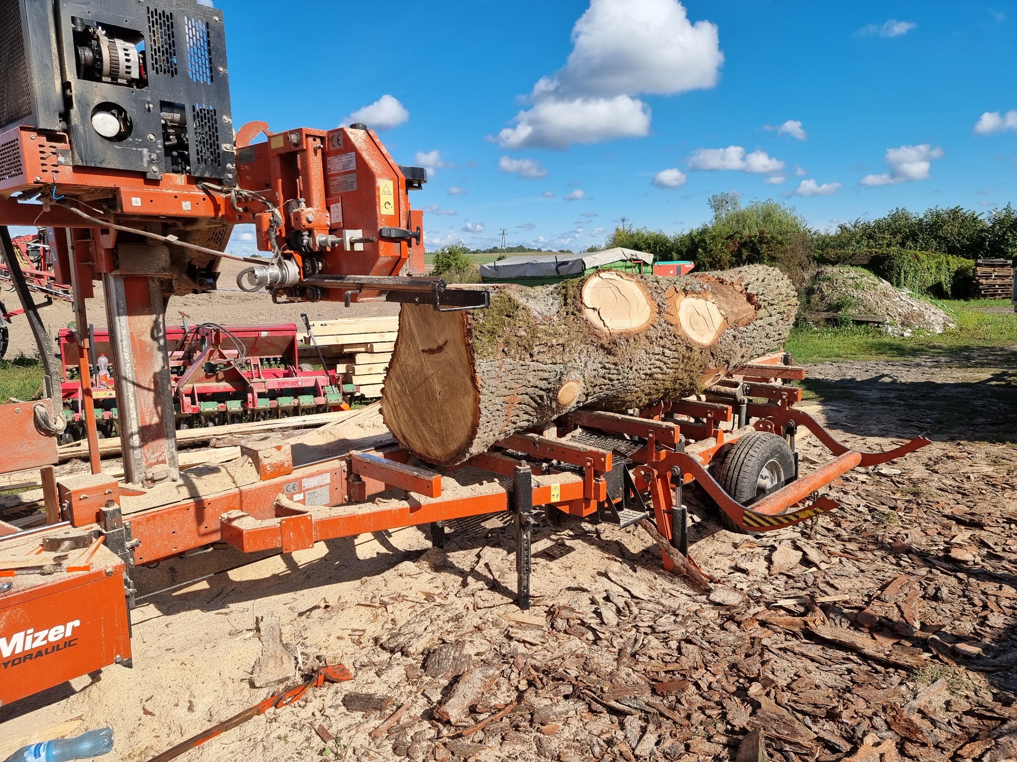 Kłoda drewna dębowa na mobilnym traku taśmowym marki Wood-Mizer, przygotowana do przetarcia na deski, widoczne świeżo ścięte słoje, w tle niebieskie niebo z chmurami i sterta kory.